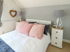 A bedroom with a bed with pink and gray pillows white bedside tables with lamps and a wooden heart decoration on the wall at Annedd Wen in Rhosneigr
