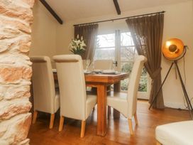 A dining room with a wooden table and white chairs next to windows with brown curtains and a standing lamp at Meadow Thatch in Cockington