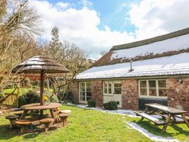 An outdoor garden area with a wooden round picnic table with stools and a thatched umbrella and a wooden bench table outside a stone cottage with a snow-covered roof at Meadow Thatch in Cockington