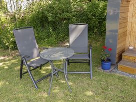 A patio area with two metal chairs and a round table on grass next to a wooden structure with steps and a blue flower pot with red flowers at Florence Pod in Pembroke