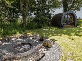 An outdoor area with a circular gravel fire pit surrounded by wooden stools and a curved wooden pod with glass doors and patio furniture at Florence Pod in Pembroke
