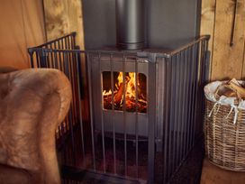 A wood burning stove with fire inside surrounded by a metal safety gate and a basket of firewood next to a brown armchair at Braeburn Lodge in Coxbridge near Glastonbury