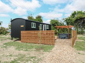 A small black wooden cabin with mint green windows and a hot tub under a wooden pergola with a fenced enclosure and wood chip path at The Heron in Lode near Bottisham