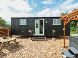 An outdoor area with a black wooden cabin with green windows and door a wooden picnic table and a hot tub under a wooden pergola at The Heron in Lode near Bottisham