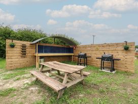 An outdoor picnic area with a wooden table two barbeques and a metal shelter against a wooden fence at The Heron in Lode near Bottisham