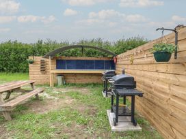 An outdoor area with two black barbecues a wooden picnic table a covered wooden bench and green plants at The Heron in Lode near Bottisham