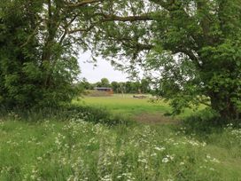 A field with green grass and wildflowers framed by two large trees with a small structure and picnic table in the distance at The Heron in Lode near Bottisham