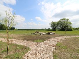 An open field with two young trees on either side of a wood chip path leading to picnic tables and logs at Barn Owl in Lode near Bottisham