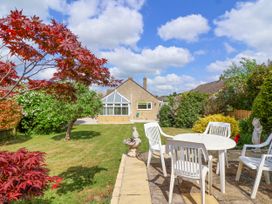 A garden with a lawn a tree red leaves a stone patio with a white table and white chairs at Hillside in Bishops Cleeve