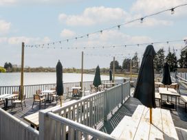 An outdoor dining area with wooden tables and chairs and closed umbrellas beside a body of water at Meadowview - Billing Aquadrome in Billing Aquadrome Holiday Park