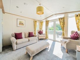 A living room with beige sofa and armchairs with red cushions yellow curtains and glass doors leading outside at Serenity Shores -  Billing Aquadrome in Billing Aquadrome Holiday Park