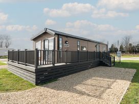 A single-story cabin with a wooden deck and railing on a gravel and grass lot at Billing Bliss - Billing Aquadrome in Billing Aquadrome Holiday Park