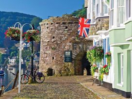 A stone archway and buildings with hanging flower baskets near a riverside with boats and hills in Dartmouth
