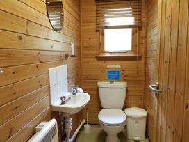 A small bathroom with a toilet, sink, mirror, window, trash bin, and wooden walls at Cedar Lodge in Rosliston