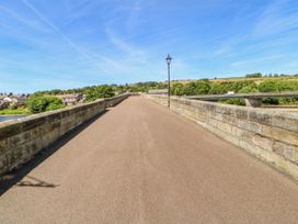 A paved pedestrian bridge with stone railings and a single lamppost over a river near houses and greenery at Shaftoe Mews in Haydon Bridge