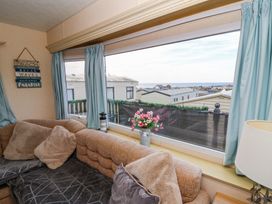 A living room with a brown couch and pillows next to a large window with blue curtains overlooking other buildings at GM18 Clarach Bay near Aberystwyth