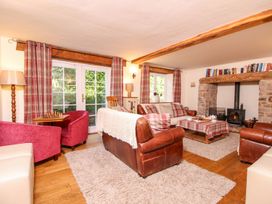 A living room with leather sofas, armchairs, a coffee table with a tablecloth, a stone fireplace, books on a wooden beam, and plaid curtains at Church Thatch in Abbotskerswell