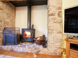 A wood burning stove with fire inside set in a stone fireplace next to a basket of wood and fireplace tools in a living room at Church Thatch in Abbotskerswell