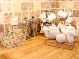 A kitchen counter with a wire basket holding an egg two French presses ceramic teapots a sugar bowl a creamer and hanging white mugs at Church Thatch in Abbotskerswell
