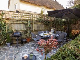 An outdoor patio with a dining table set with plates and drinks surrounded by chairs a barbecue grill and a large umbrella with string lights at Church Thatch in Abbotskerswell