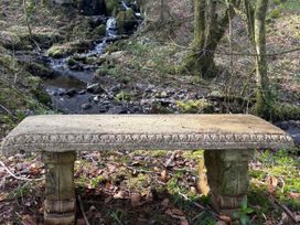 A bench near a stream with trees in the background at Bracken Ground Coniston