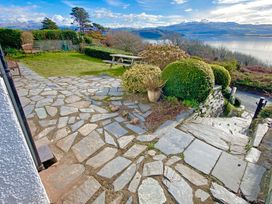 An outdoor stone patio with benches picnic table bushes and a view of water and mountains at Bryn Eithin in Borth-y-Gest near Porthmadog