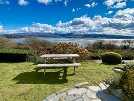 A picnic table on grass with a stone pathway and bushes near a body of water and hills at Bryn Eithin in Borth-y-Gest near Porthmadog