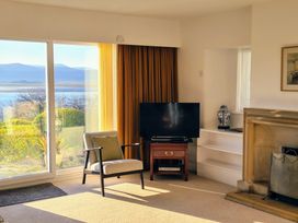 A living room with a chair a television on a wooden stand a fireplace and large windows showing an outdoor garden and mountains at Bryn Eithin in Borth-y-Gest near Porthmadog
