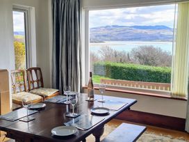 A dining area with a wooden table set with plates and wine glasses near a large window overlooking a garden hedge and distant hills at Bryn Eithin in Borth-y-Gest near Porthmadog