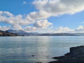 A coastal view with calm water, rocky shoreline, hills in the distance, and a cloudy sky at Bryn Eithin in Borth-y-Gest near Porthmadog