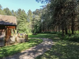 A wooden cabin with a porch and grill on a grassy area next to a gravel path surrounded by trees at Birch Lodge in Rosliston