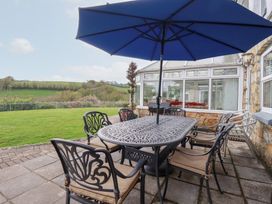 An outdoor patio area with a metal dining table and chairs under a large blue umbrella beside a house with glass conservatory and green lawn at Chapel Green in Polgooth