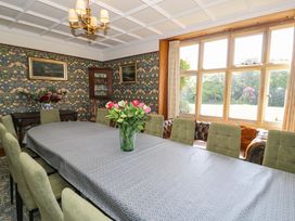 A dining room with a long table covered with a tablecloth and chairs around it with a vase of flowers on the table large windows and patterned wallpaper at Plas Gwynfryn in Llanbedr