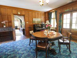 A room with wooden panel walls a blue patterned rug an octagonal wooden table with chairs a piano and floral curtains at Plas Gwynfryn in Llanbedr