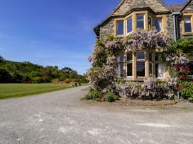 A stone house with flowering vines around windows and a gravel driveway with grass and trees at Plas Gwynfryn in Llanbedr