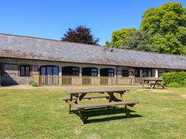 A grassy outdoor area with wooden picnic tables in front of a long stone building with black windows and a wooden fence at Keepers Cottage in Winterborne Stickland