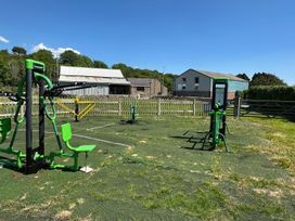 An outdoor exercise area with green fitness equipment and a fenced yard with buildings in the background at The Mill in Abererch near Pwllheli