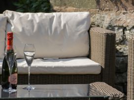 An outdoor wicker sofa with white cushions beside a glass table holding a bottle of cava and two empty glasses at Trevor Cottage in Llandudno