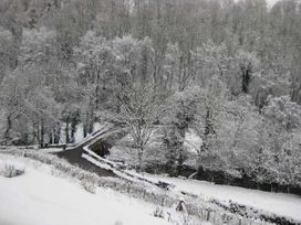 A snowy landscape with a road crossing a stone bridge and trees covered in snow at Little Cow House in Pontfaen near Chirk