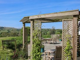 A wooden pergola with climbing plants and wooden outdoor furniture on a paved patio with a view of green hills and blue sky at The Old Beams at Pont y Forwyn in Llansantffraidd-ym-Mechain near Welshpool