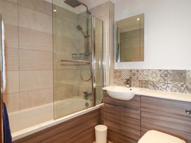 A bathroom with a bathtub and shower, a sink with a mirror above it, patterned tiles on the wall, and wooden cabinets at Pink House in Mevagissey