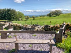 An outdoor seating area with wooden benches around a stone fire pit overlooking green fields and trees at Pant Dafydd Goch in Llangollen