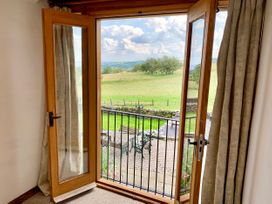 A pair of wooden framed glass doors open to a balcony with metal railing overlooking a patio with benches and a table and a grassy field with trees at Pant Dafydd Goch in Llangollen