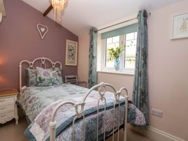 A bedroom with a bed floral bedding and cushions a bedside table with lamp and a window with curtains and a vase of flowers at Puffitts Cottage in Bourton-on-the-Water