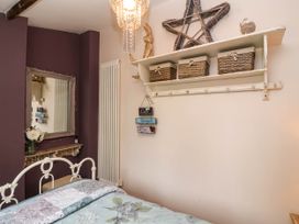 A bedroom with a white metal bed frame a quilted bedspread a wall shelf with baskets and decorations and a mirror on a purple wall at Puffitts Cottage in Bourton-on-the-Water