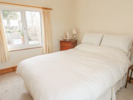 A bedroom with a bed covered in white bedding next to a window with cream curtains and a wooden nightstand with a lamp at Jasmine Cottage in Rhuddlan