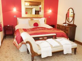 A bedroom with a large bed with red bedding and pillows two nightstands a wooden dresser with an oval mirror and a bench with folded towels at Gelli Glud in Eglwysbach