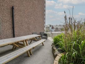 An outdoor patio area with a wooden picnic table and benches two black wicker chairs a small garden bed with plants and a stone border a wall and rooftops of nearby buildings in view at Eversley in Llandudno