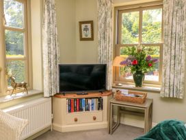 A corner with a TV on a wooden cabinet with books below next to windows with floral curtains and a vase of flowers Garden Cottage Black Bourton near Burford