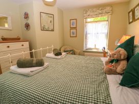 A bedroom with a green checkered bedspread a teddy bear on the bed a wicker chair a chest of drawers and a window with curtains at Garden Cottage in Black Bourton near Burford